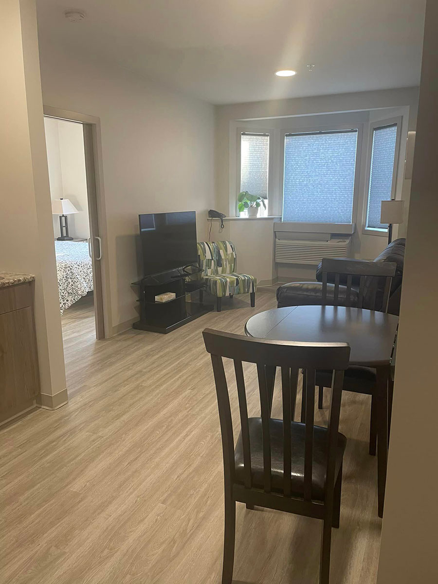 Guest suite at Kinsmen Kourts II, featuring a living/dining area with television, chair, two-seat table, fridge, and microwave.